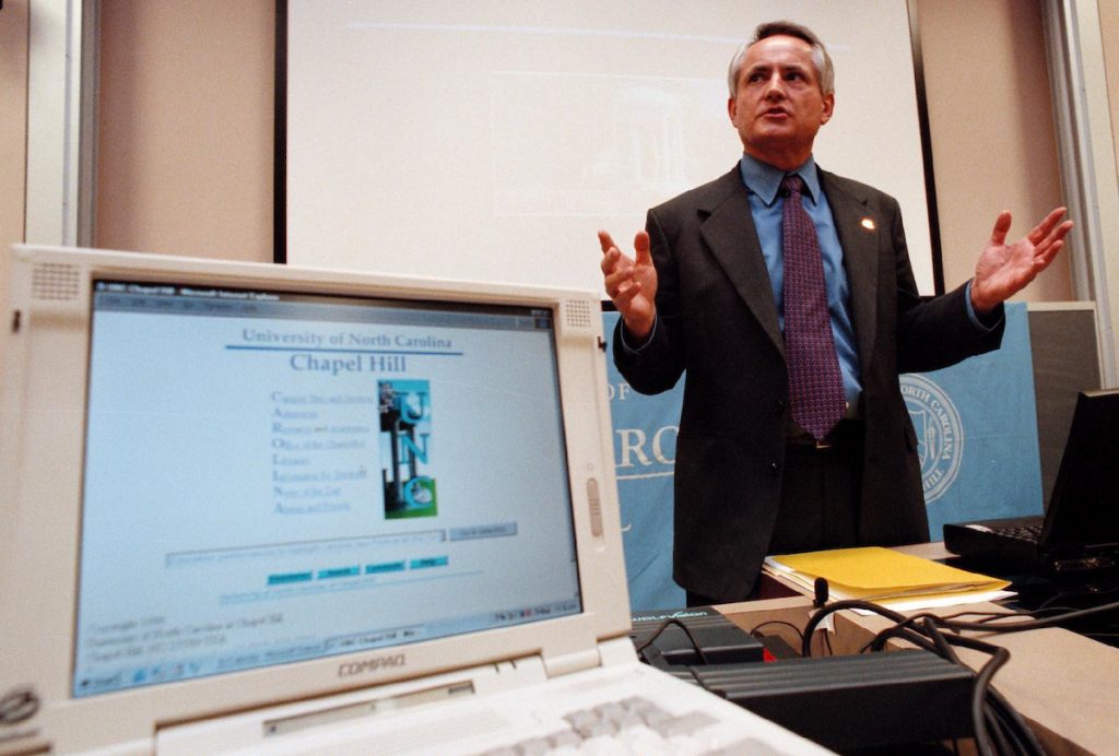 Close view of chancellor Michael Hooker speaking at press conference. In the foreground, a 1998 laptop displays the UNC webpage